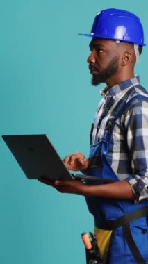 Vertical video: Modern constructor using laptop to find inspiration for building project, browsing online and making calculations or measurements on camera. Construction worker holding wireless pc to