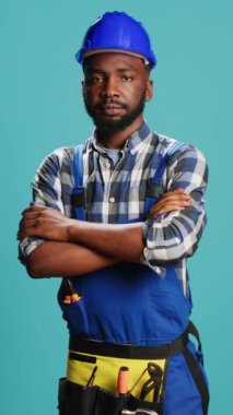 Vertical video: Cheerful smiling handyman standing over blue backdrop, wearing construction uniform and protective helmet. Professional carpenter repairman posing in studio, mechanical reconstruction.