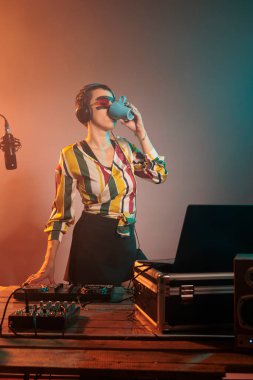 Young woman DJ drinking coffee from mug before mixing techno music, preparing to do performance on turntables. Holding caffeine drink in cup and doing remix with volume mixer.