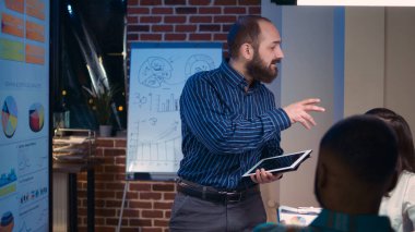 Şirket finans analisti, birlikte çalışma alanındaki iş toplantısında elinde dijital tabletle konuşuyor. Ofis çalışanları beyin fırtınası yapıyor, gelir istatistiklerini tartışıyor, ekrandaki grafiklere bakıyor.