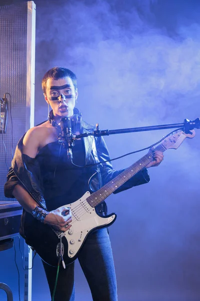 Punk rocker with cool make up singing alternative rock, playing guitar and doing concert show in studio. Ecstatic talented guitarist performing heavy metal music live with microphone and instrument.