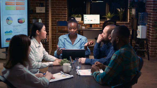 Diverse coworkers brainstorming, listening to executive in business meeting, discussing presentation. African american businesswoman talking, team leader speaking in office at night time