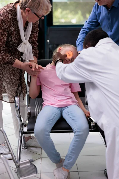 Practitioner doctor shining light into little girl patient eye while testing pupil reflexes during checkup visit consultation in hospital waiting room. Unconscious kid fainting on chair