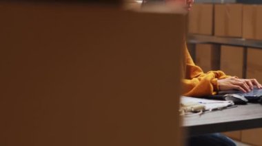 Female entrepreneur taking notes on papers and pc, working in warehouse to do inventory and shipping logistics. Young adult checking products in boxes before distribution. Handheld shot.