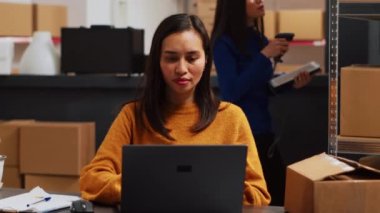 Asian employee working on stock logistics with laptop, planning order shipment for business development in warehouse. Woman doing supply chain management at desk, retail goods. Handheld shot.