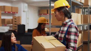 Asian guy organizing boxes with supplies on shelves, doing quality control with female entrepreneur in warehouse. Employee putting packages on racks, preparing for order management.