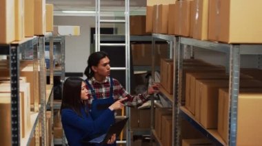 Warehouse employees working on inventory with laptop, walking around storage room racks with carton boxes. Entrepreneur and worker planning stock distribution, small business concept.