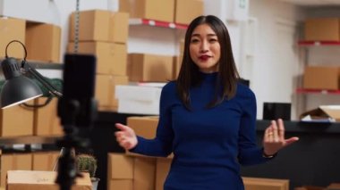 Young entrepreneur filming teleshopping video on phone, advertising products in warehouse space. Female worker using social media channel to create online ad promotion. Handheld shot.