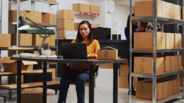 Young startup owner working on laptop to plan logistics, preparing products order to ship to clients. Female employee packing merchandise in boxes, taking notes for storage inventory.