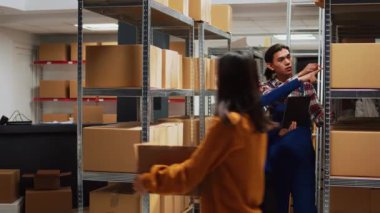 Team of people checking stock supplies on laptop, working with merchandise boxes in warehouse space. Man and woman doing teamwork for inventory and products logistics in storage room.