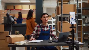 Asian worker recording ad video in storage room, using social media streaming platform to advertise small business in warehouse. Group of people doing teamwork for online promotion.
