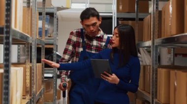 Asian man in crutches working with entrepreneur in depot, looking at inventory information on tablet. Male worker and small business owner planning products distribution. Handheld shot.