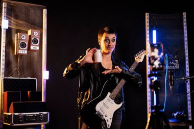 Woman rocker holding bottle of pills reviewing health product in front of camera while recording new rock song using electric guitar in sound studio. Musician working at grunge album practicing songs