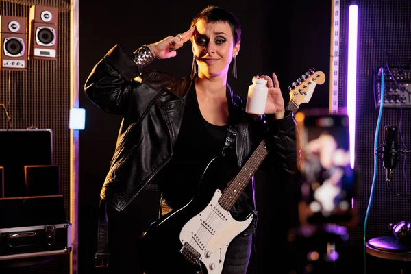 Woman musician holding bottle of pills showing medicament at camera during music lesson in sound studio, reviewing health product. Rockstar playing at electric guitar working at new heavy metal song