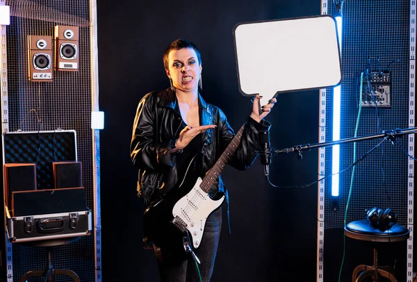 Woman musician holding blank white cardboard for advertise text while wearing electric guitar preparing to play rock song in studio. Guitarist using electric instrument to perform heavy metal music