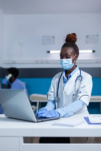 病院の医療専門医は ウイルス保護マスクを着用しながら コンピュータを使用して病気の患者データベースを更新します 専門医が患者の病歴を記入し治療結果を調べる — ストック写真