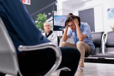 Exhausted young patient having medical consultation waiting for doctor to come to discuss healthcare treatment during checkup visit in hospital area. Tired asian man having health probleme.