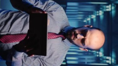 Vertical video: Male infrastructure specialist using tablet for hardware digitalization, inspecting cloud computing and cluster computer. Data center technician running server diagnostics. Tripod shot