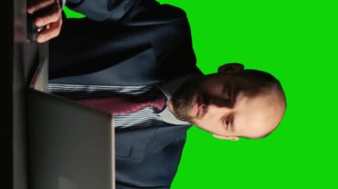 Vertical video: Entrepreneur looking at data on isolated greenscreen, and working on laptop. Young businessman in suit and tie standing over blank mockup and chroma key background, white collar.
