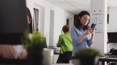 Female employee browsing internet on smartphone, working in startup office. Young business worker using mobile phone to analyze statistics report for company development. Handheld shot.