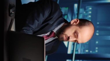 Vertical video: Male infrastructure engineer working in render farm running diagnostics on mainframe computer system in modern data center. Database admin controlling work of server cabinets on laptop