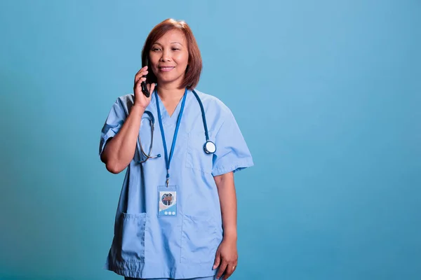 Specialist nurse talking at phone with remote doctor discussing medical treatment during appointment in studio with blue background. Medical assistant wearing coat explaining patient insurance