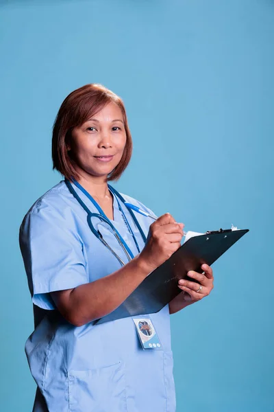 Medical assistant checking disease expertise writing medication treatment on clipboard during checkup visit. Physician nurse with blue uniform and stethoscope working in health care industry
