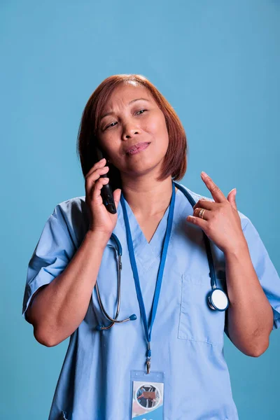 Practitioner nurse talking at smartphone with remote doctor discussing medical treatment during appointment in studio with blue background. Medical assistant wearing coat explaining patient insurance