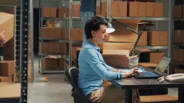 Entrepreneur using laptop to plan storage logistics, preparing merchandise order to ship to customers. Young employee putting products in packages, taking notes for stock inventory.