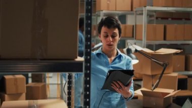 Young entrepreneur using scanner and tablet to do inventory, scanning bar code from packages on warehouse shelves. Woman checking stock of merchandise in boxes, supply chain logistics.