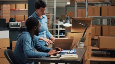Young partners checking quality of merchandise packed in cardboard boxes, preparing order to ship to customers. Team of employees working on shipping products from warehouse shelves.