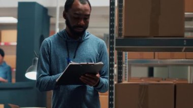 Small business partner working on merchandise inventory, taking notes on clipboard papers. Male worker checking products in cardboard boxes, looking at stock on warehouse shelves. Handheld shot.