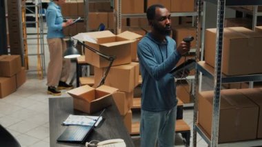 Small business owner scanning bar code on carton boxes, working on doing merchandise inventory in warehouse space. Male worker taking packages from sorage room racks and checking them.