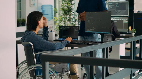 App developer talking to coder with chronic impairment in agency office, explaining source code programming on computer. Wheelchair user coding script with new user interface.