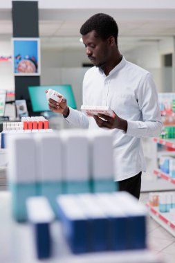 Young african american taking cardiovascular supplements from drugstore shelf, holding two heart medication packages, checking instruction. Customer buying medicaments, choosing prescription treatment