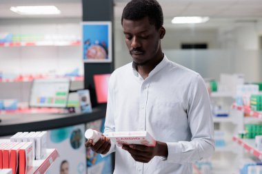 Young african american man choosing cardiovascular supplements in drugstore, holding two heart medication packages, reading instruction. Buyer thinking, buying medicaments, prescription treatment