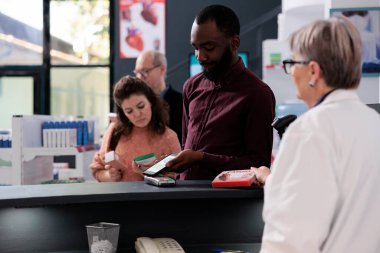Young adult client putting smartphone on pos terminal paying for supplements in pharmacy after discussing health care treatment with pharmacist. Client at drugstore counter buying prescription pills