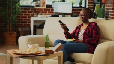 Relaxed person laughing at comedy movie on tv and drinking alcohol, holding bowl of snacks in living room. Young woman enjoying film on television with chips and beer, takeout food.