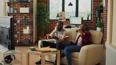 African american people eating slices of pizza in front of tv, binge watching favorite show at home. Relaxed modern couple serving fast food meal and drinking bottles of beer.