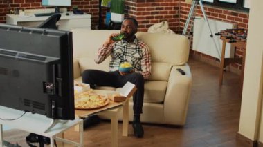 African american adult drinking beer and eating chips, enjoying comedy tv show in living room. Modern man having fun with movie on television, serving takeaway fast food at home.