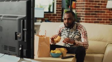African american guy having fun eating burgers and drinking beer, watching comedy movie on television. Male adult enjoying fast food delivery and alcohol, watch tv show in living room.