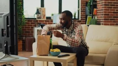 Cheerful guy eating cheeseburger with fries at home, enjoying fast food meal order from takeout and watching comedy movie on tv. Young man serving burger and drinking beer bottle.