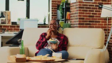 African american woman eating bowl of chips on sofa, laughing at comedy tv show in living room. Young adult feeling cheerful and relaxing at home with snacks and beer. Tripod shot.