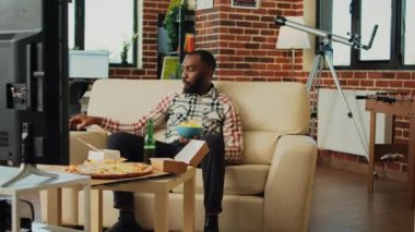 Young person drinking bottle of beer and eating snacks, enjoying favorite tv show in living room. Cheerful guy having fun with film on television, serving takeaway burgers, pizza and noodles.