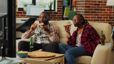 African american partners eating pizza slices at home, feeling happy watching film together. Relaxed couple having fun eating takeaway delivery food in living room, leisure activity.