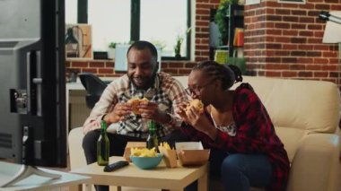 Modern life partners taking bite of cheeseburger, eating fast food from delivery takeaway at home. Young couple having dinner and enjoying comedy movie on television channel, beer bottles.