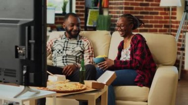 Happy life partners binge watching tv show together, holding bottle of beer and eating chips at home. Young people in relationship feeling relaxed with movie on tv, eating snacks.