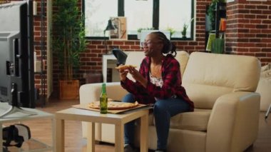 African american woman watching tv and eating pizza, laughing at comedy movie in living room. Young person serving fast food delivery meal and binge watching favorite tv show.