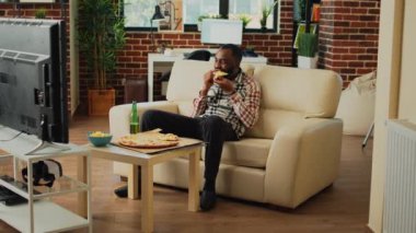 Cheerful adult taking bite of pizza at home, eating takeaway food delivery and drinking alcohol. Young relaxed man having fun watching action movie on television, eating snacks.
