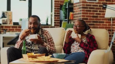 African american people eating noodles with chopsticks and binge watching action tv series at home. Young partners feeling relaxed eating asian takeaway food, watch film. Tripod shot.